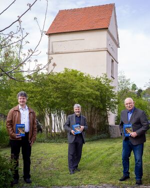 Martin Lauber, Heimatverein Untergrombach, Autor Thomas Adam und Wolfram von Müller, Heimatverein Obergrombach (v.l.n.r.), mit der neu erschienenen Veröffentlichung „Strom für das Grombachtal“. Die beiden Heimatvereine Ober- und Untergrombach haben die Schrift gemeinsam herausgegeben. Die Ursprünge der Transformatorenstation in der Obergrombacher Hauptstraße (Hintergrund) gehen auf die Anfänge der regionalen Stromversorgung vor hundert Jahren zurück.